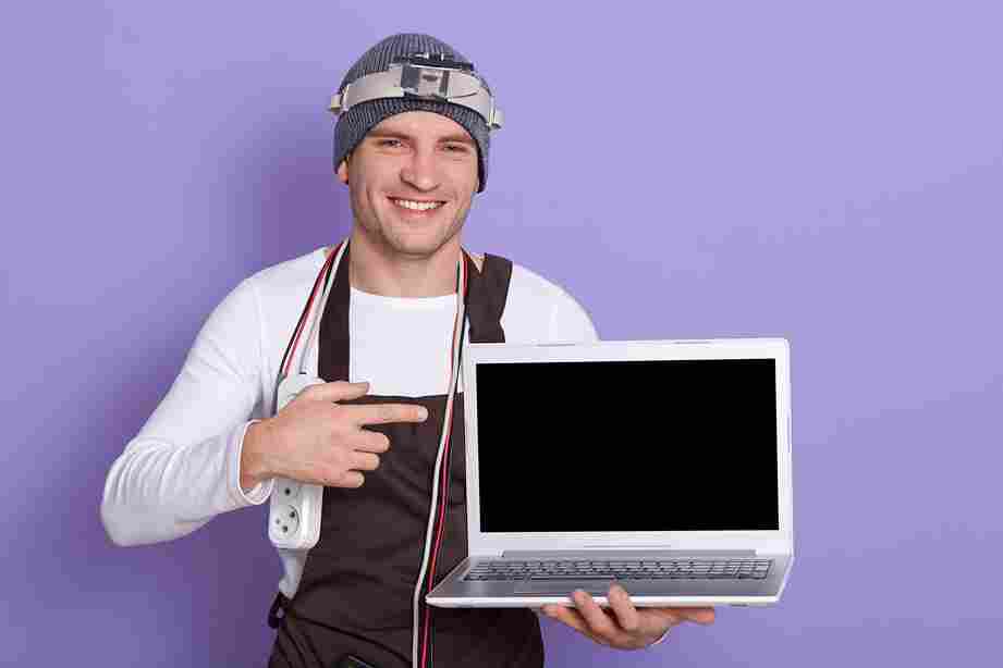 Cheerful technician holding laptop with blank screen, showing finger and wearing cords and adapter for laptop repair.